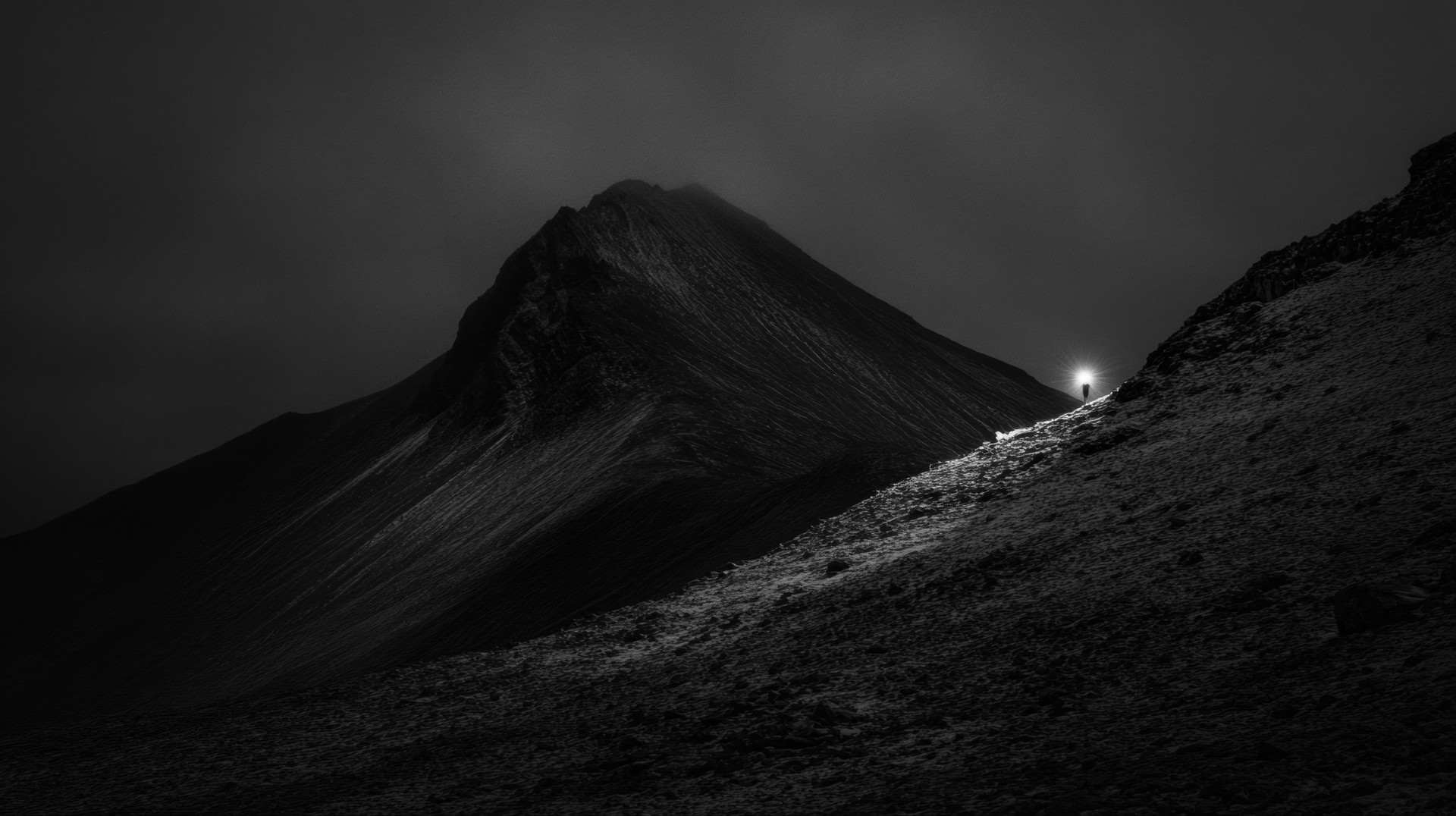 Schwarz-weiß Foto von einem Berg bei Nacht. Eine einzelne Person hält eine leuchtende Laterne und steht auf einem schneebedeckten Hang vor einem dunklen Gipfel.