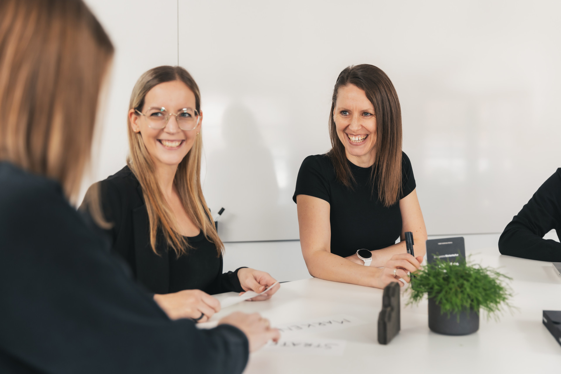 Drei Frauen sitzen lächelnd an einem Tisch in einem Büro. Zwei schauen zur Kamera. Auf dem Tisch liegen Papiere und steht eine kleine Pflanze.